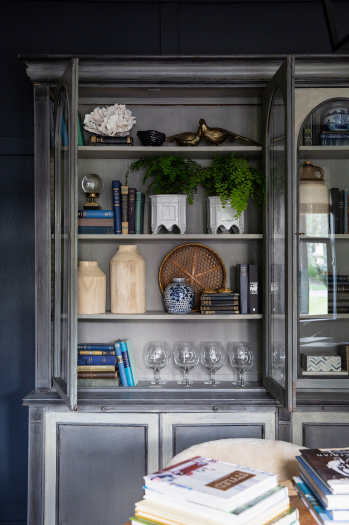 A styled gray wood cabinet with glass doors displaying layered decor, including stacked books, white ceramic planters with green ferns, carved wood vessels, a woven tray, blue-and-white pottery, brass bird figurines, and a row of etched glassware on the bottom shelf.