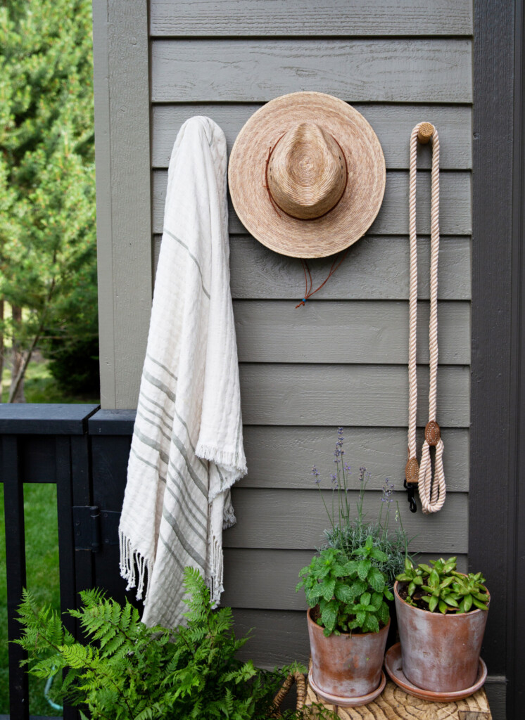Outdoor wall with striped neutral towel, woven straw hat, and rope leash hanging on hooks, styled above terracotta potted plants and greenery