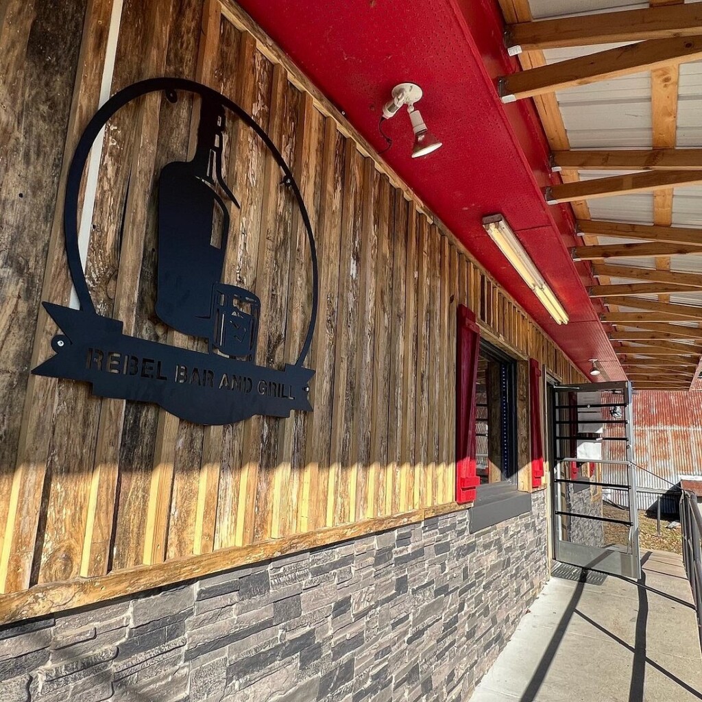 Exterior of Rebel Bar and Grill featuring rustic wood siding, a stone base, and bold red trim along the roofline. A black metal sign with a train silhouette is mounted on the wall near the entrance, with sunlight casting strong shadows along the walkway.