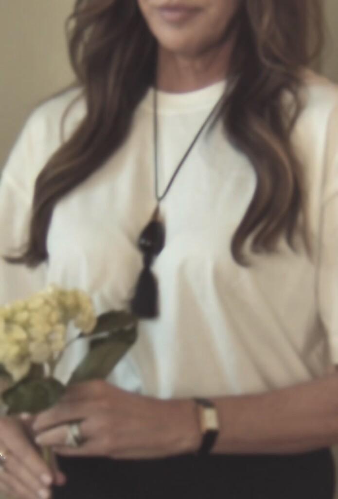 A softly lit, slightly blurred image of a woman wearing a simple white top, styled with a long black tassel necklace. She holds a small bouquet of light-colored flowers, with neutral tones and a warm, understated, minimal aesthetic.