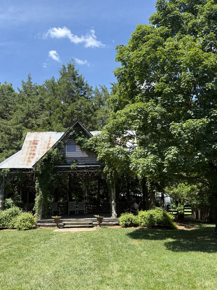 Charming rustic cottage with a weathered metal roof, shaded porch, and lush greenery surrounding a manicured lawn on a bright day.