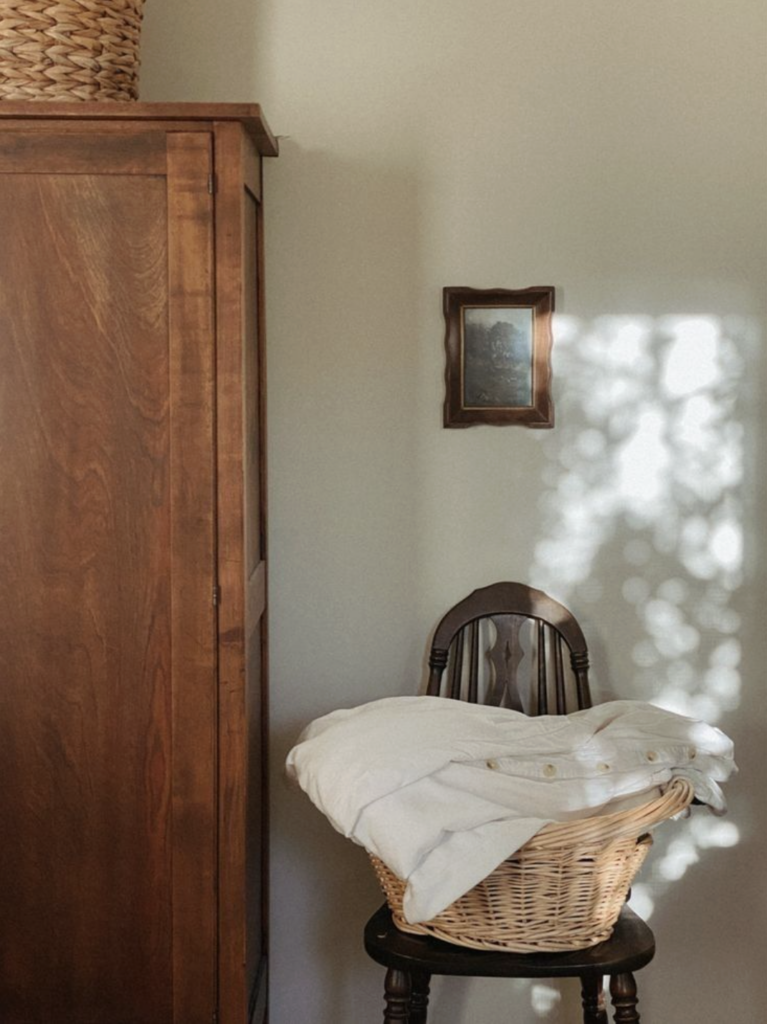 Warm, minimal interior corner with a wooden cabinet, small framed artwork, and a wicker basket filled with linens on a chair in soft natural light.