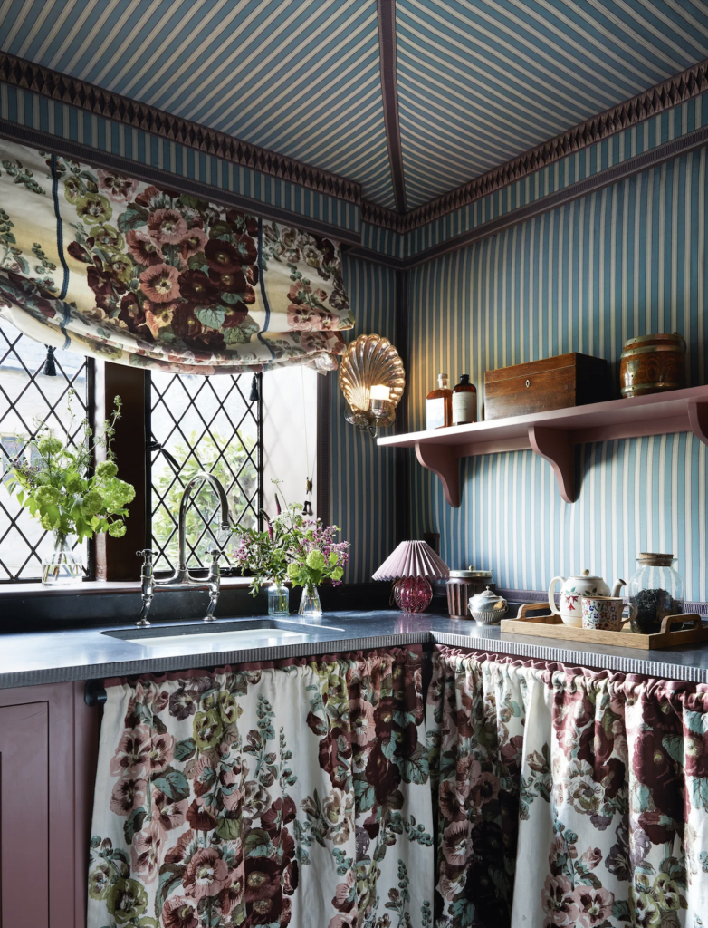 Charming English-style kitchen with blue striped walls and ceiling, a floral Roman shade and sink skirt, brass shell sconce, and layered vintage decor for a cozy, collected look.