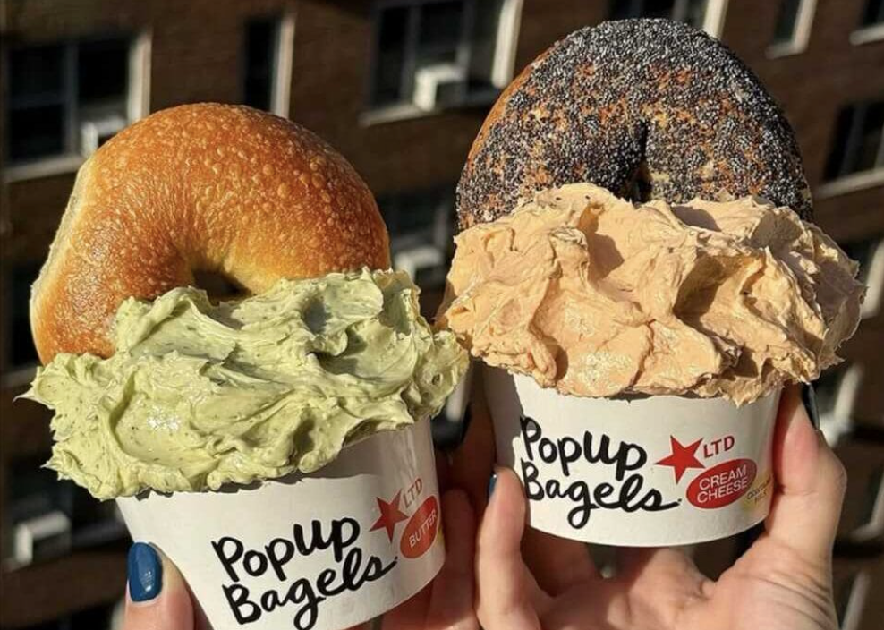 Two hands holding oversized bagels in branded “Popup Bagels” cups, each piled high with whipped cream cheese—one plain bagel with green spread and one poppy seed bagel with a tan spread—shot outdoors in warm sunlight.
