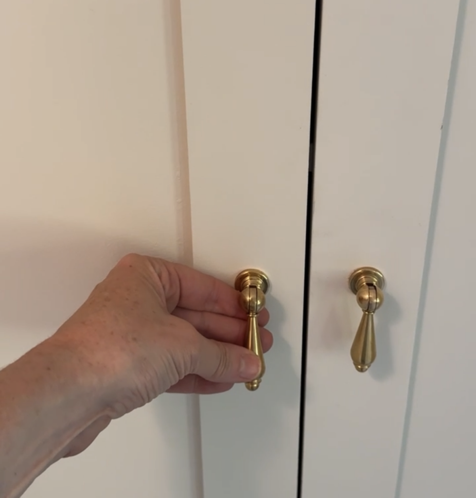 A close-up of a hand adjusting a polished brass teardrop cabinet pull on a white paneled cabinet door, showing how the hardware sits tightly in place without swinging freely.