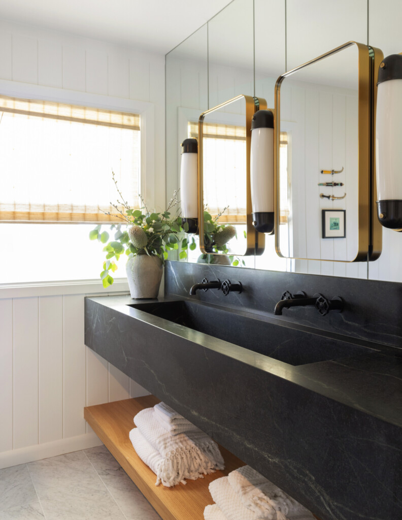 Modern bathroom vanity with black stone sink, brass mirrors, and neatly folded neutral towels