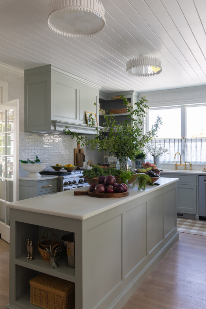 A light-filled kitchen with soft sage cabinetry, a large paneled island, white countertops, and brass fixtures, styled with fresh greenery and produce for a layered, inviting, and lived-in feel.