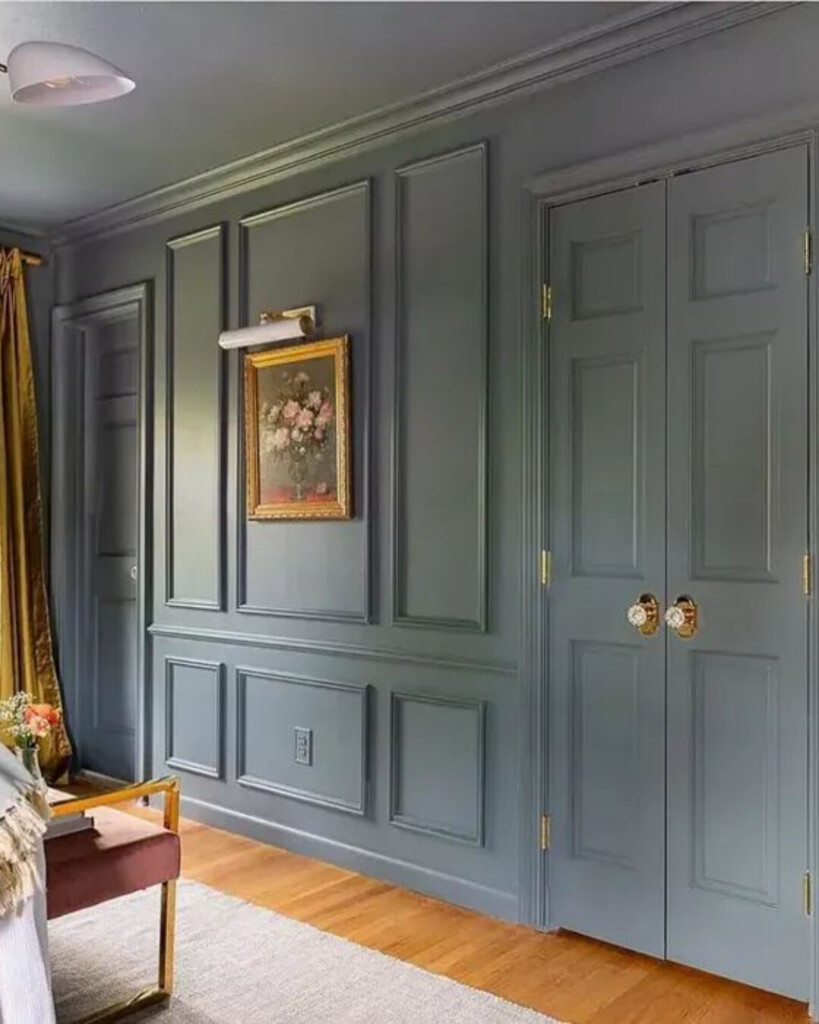 A paneled room painted in a muted blue-gray with detailed wall molding, brass hardware, warm wood floors, and a traditional framed artwork creating a layered, classic feel.