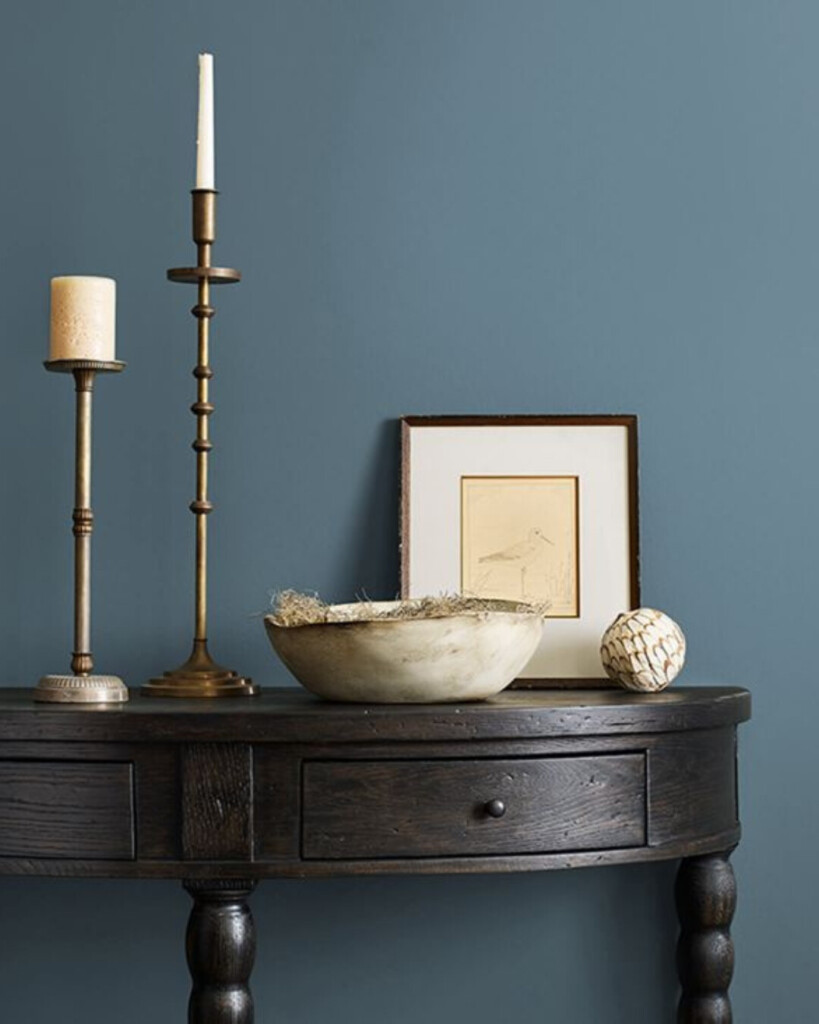 A styled vignette with a deep dusty blue wall, dark wood console table, brass candlesticks, and neutral decor highlighting the richness of the paint color.