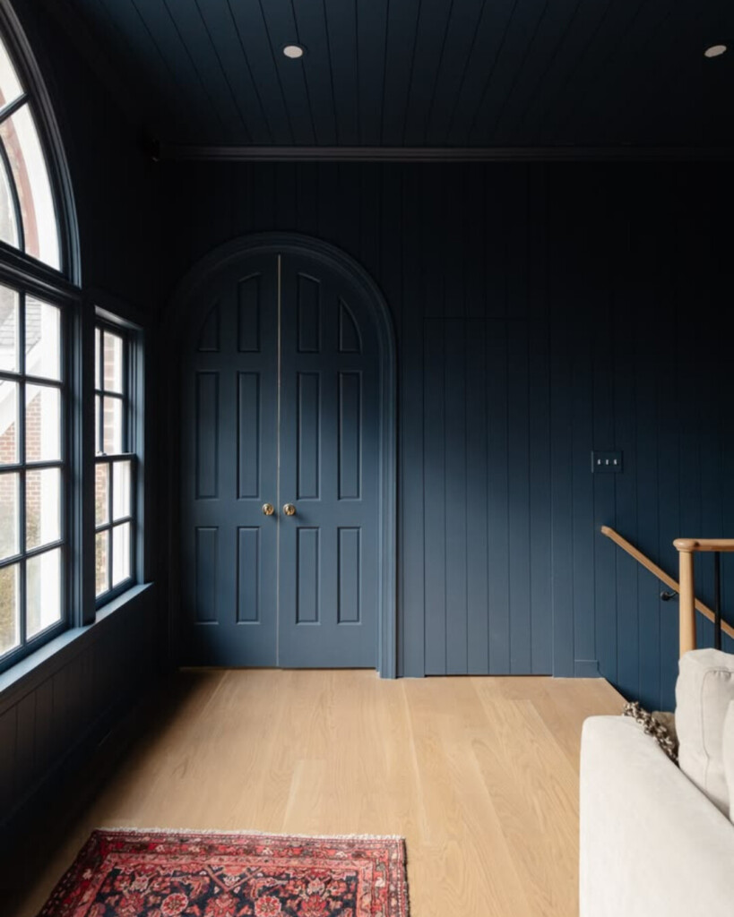 A dark, moody blue hallway with paneled walls, arched doorway, and natural light creating contrast against the deep color.
