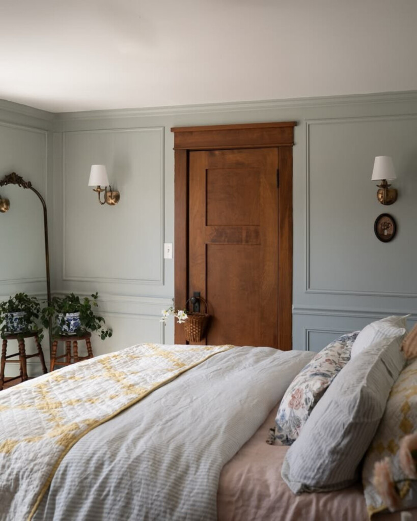 A soft, pale blue bedroom with classic wall molding, warm wood door, brass sconces, and layered neutral bedding for a calm, timeless feel.