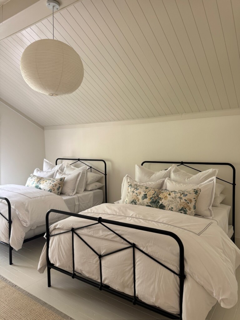 Light, airy bedroom with two black metal twin beds dressed in crisp white bedding and floral accent pillows, set beneath a sloped beadboard ceiling with a large paper lantern pendant and soft neutral tones throughout.