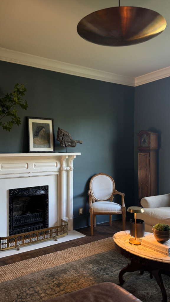 A moody, refined living room with deep blue-green walls, a classic white fireplace mantel, antique wood furniture, and warm brass accents, layered with soft textiles and vintage artwork for a timeless, collected feel.