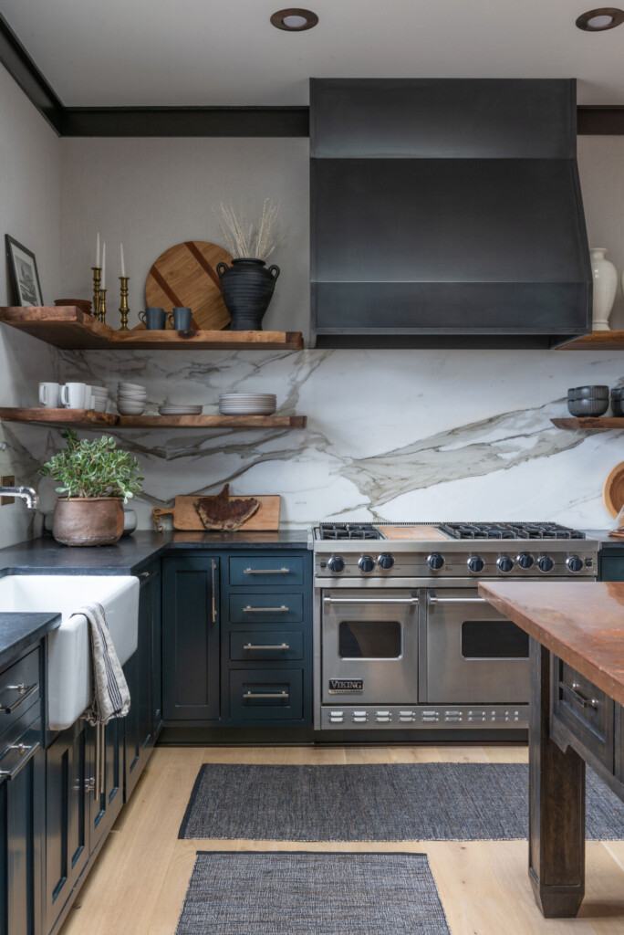 A tailored kitchen with deep blue cabinetry, a dramatic marble slab backsplash, warm wood shelving, and a large stainless steel range, layered with natural textures and brass accents for a refined, functional space.